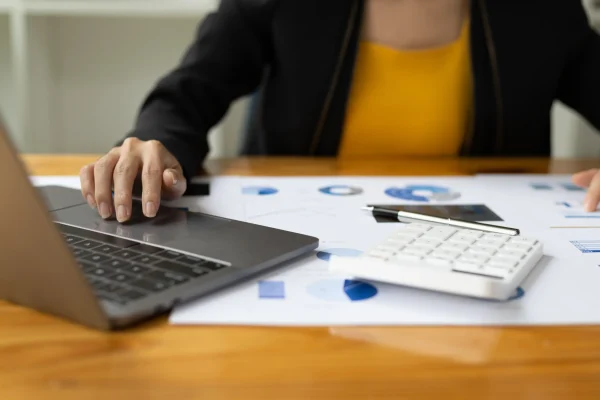 Mujer usando su laptop con CONTPAQi® Factura Electrónica, acompañadas por una calculadora y documentos analíticos. Profesional contable utilizando CONTPAQi® Factura Electrónica para procesar información financiera en una computadora.