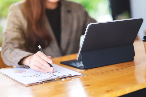 Supervisora usa una tablet y escribe resultados para evaluar el rendimiento en la gestión de producción de su área de trabajo. Ejecutiva analiza gráficas y toma notas como parte de la gestión de producción, combinando tecnología y planeación estratégica.