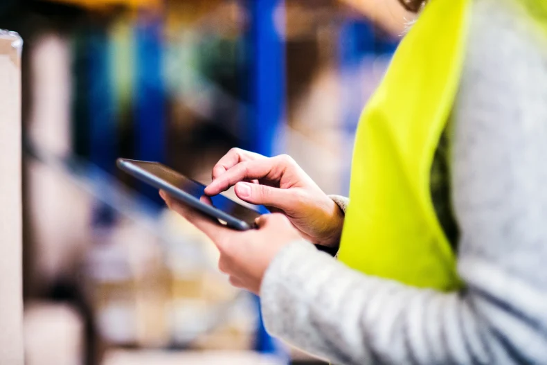 Persona con chaleco de seguridad usando una tablet en un almacén, gestionando el sistema de control de inventarios.