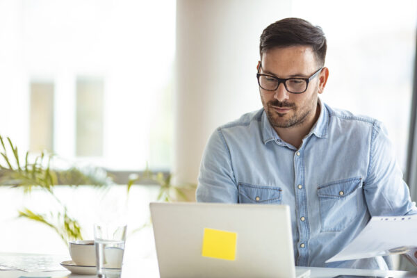 Profesional revisa documentos físicos y digitales gracias a su sistema de facturación electrónica conectado en línea. Hombre compara información impresa con datos en su computadora usando un sistema de facturación electrónica eficiente.
