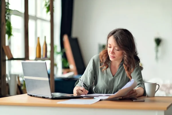 Jornada laboral en la que una profesional lleva a cabo la gestión de nómina, incluyendo el registro de incidencias o la validación de recibos. Una persona toma notas mientras revisa información en su computadora portátil, para organizar información relacionada con la gestión de nómina.