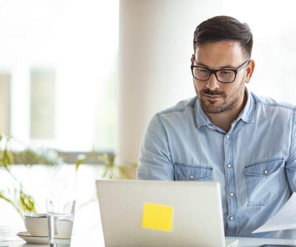 Profesional revisando archivos para la gestión de nómina en su computadora. Colaborador revisa simultáneamente archivos físicos y digitales. Una práctica durante procesos de gestión de nómina.