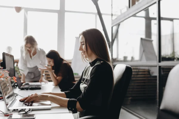 Sentada frente a una laptop, una usuaria interactúa con distintos dispositivos en una jornada laboral, representando la flexibilidad de acceso a sistemas como CONTPAQi® Kursa. Mujer trabaja en su laptop en un espacio de oficina compartida, mientras consulta información de CONTPAQi® Kursa para aprendizaje en línea.