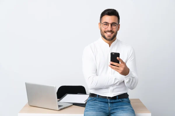 En un entorno blanco y minimalista, un joven empresario revisa su teléfono móvil, posiblemente accediendo a sistemas como Contpaqi nube desde su dispositivo. Un hombre sonriente, vestido de manera formal, observa su celular con una laptop al fondo; compatible con Contpaqi nube.