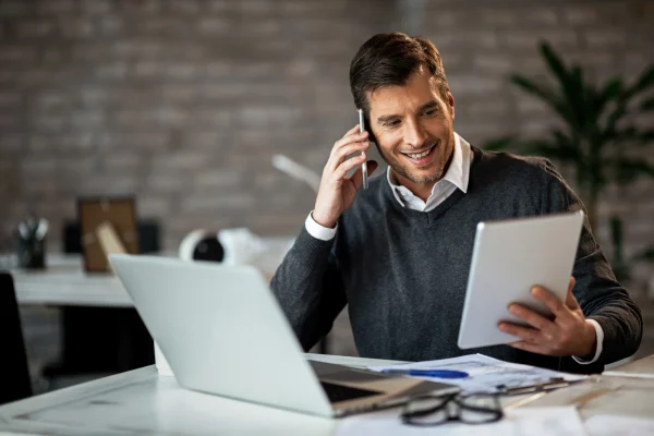 Hombre trabajando con laptop y tablet mientras realiza una llamada telefónica, en un entorno de oficina donde utiliza CONTPAQi Contabiliza®. Usuario revisando información contable con CONTPAQi Contabiliza®, accediendo desde diferentes dispositivos gracias a su sistema en la nube.
