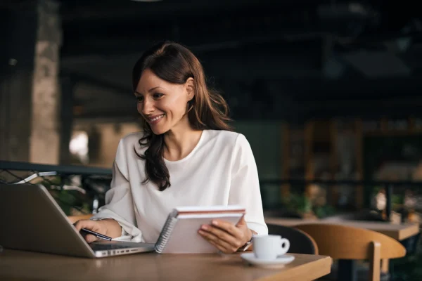 Persona trabajando desde laptop con libreta en mano, utilizando el sistema contable CONTPAQi Contabiliza® para organización financiera. Mujer analizando información en laptop y tomando notas con ayuda de CONTPAQi Contabiliza® en un espacio abierto.