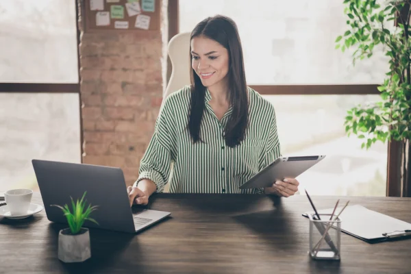 Usuaria revisando archivos impresos y digitales simultáneamente, una práctica habitual en procesos de emisión y control fiscal con CONTPAQi® Timbra. Una mujer consulta documentos físicos mientras trabaja con su laptop en una oficina, a través del uso de CONTPAQi® Timbra.