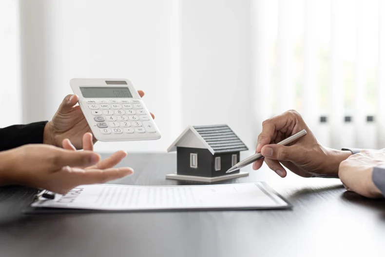 Una persona sostiene una calculadora mientras otra señala documentos junto a una maqueta de vivienda, un reflejo de los ajustes financieros que plantea la Reforma Infonavit 2025.