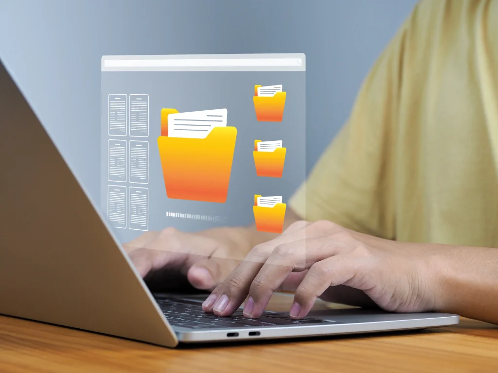Archivos digitales accesibles al escribir en la computadora, ejemplo de soluciones de respaldo de datos. Carpetas amarillas flotan mientras alguien usa una laptop, relacionadas con soluciones de respaldo de datos.