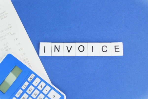 Bloques de madera que forman la palabra "INVOICE" junto a una calculadora azul y un recibo sobre un fondo azul.