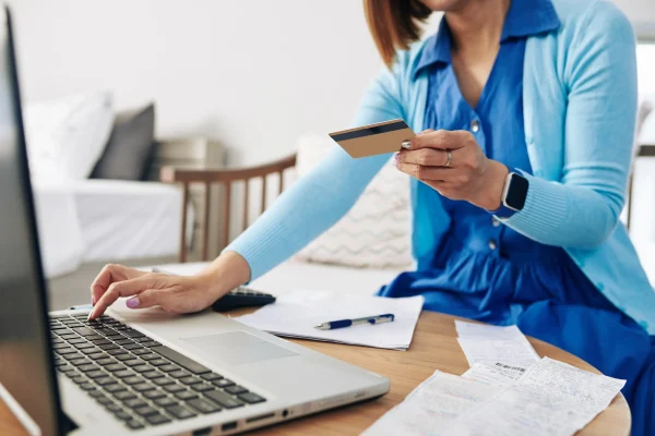 Cómo cumplir con los requisitos del SAT para facturar en línea desde casa. Mujer sentada frente a una laptop sosteniendo una tarjeta de crédito y revisando recibos impresos para facturación.