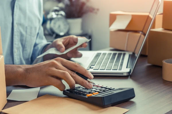 requisitos del SAT para facturar en línea_3 Persona utilizando una calculadora y revisando tickets de compra frente a una computadora portátil con paquetes de envío al fondo.